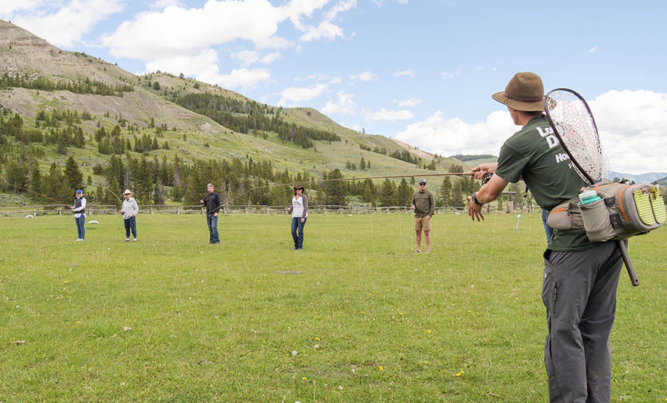A group of people stands spaced out in a grassy field with mountains and trees in the background. In the foreground, at a Montana Dude Ranch, a person wearing a hat and carrying fishing gear appears to be instructing or demonstrating something to the group. The sky is partly cloudy.