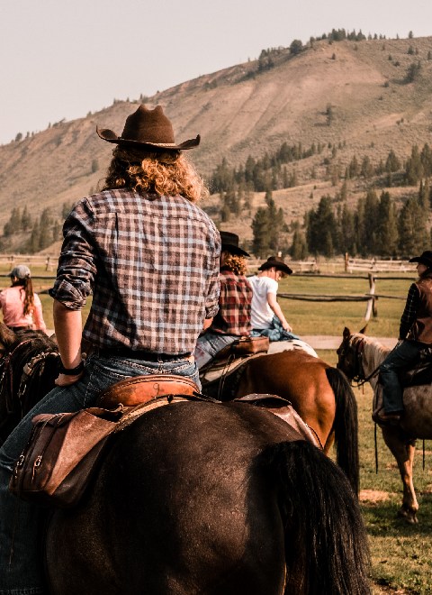 thumb-horseback-program-learn-to-ride-yellowstone-horseback-riding-opt2 (2) A group of individuals wearing cowboy hats riding horses across a vast, open field under a clear blue sky.