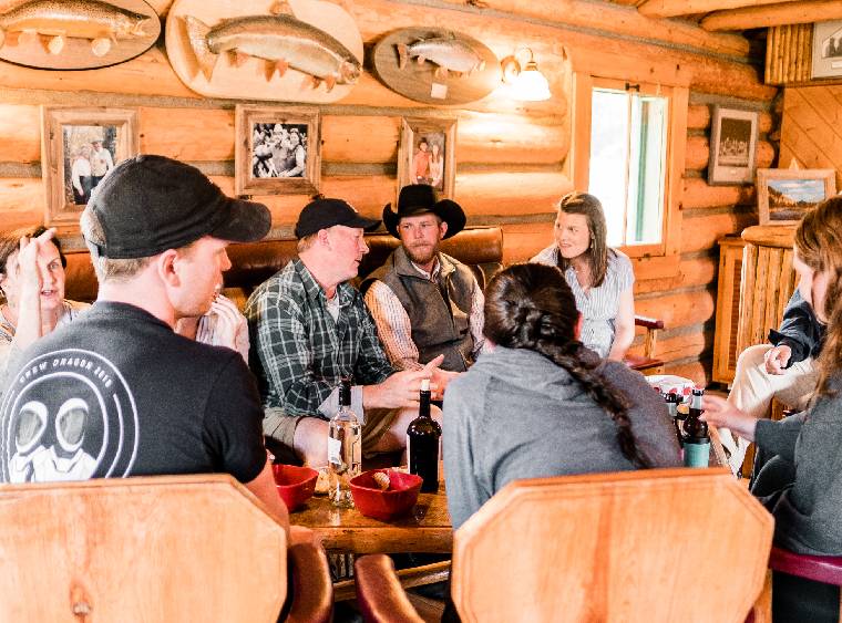 thumb-loding-happy-hour-lodging-near-yellowstone-2 (2) A group of individuals gathered around a table inside a cozy log cabin, engaged in conversation and sharing a meal.