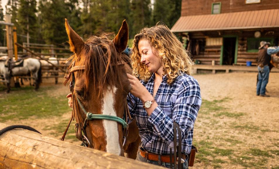thumb-specialty-weeks-first-section-montana-dude-ranch-opt A person with curly hair, wearing a plaid shirt, is petting a brown horse with a white stripe on its face. They are outdoors next to a wooden fence, with a rustic building and other horses in the background. The setting appears to be a Montana Dude Ranch.