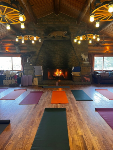 A cozy yoga studio with wooden floors and log cabin-style interior features a large stone fireplace with a fire burning. Yoga mats of various colors are laid out in rows on the floor. The room is warmly lit with rustic hanging lights, and large windows let in natural light.