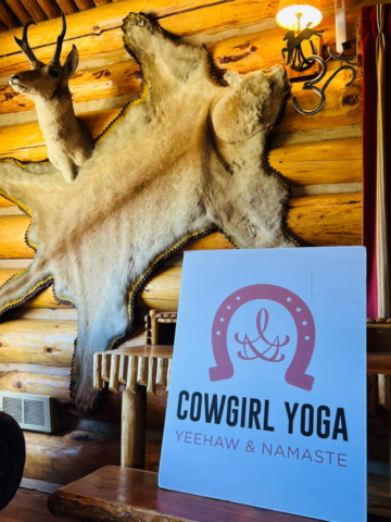 A wooden room with mounted animal hide and antelope head on the wall. A sign in the foreground reads "COWGIRL YOGA YEEHAW & NAMASTE" with a red horseshoe and lasso graphic. Warm lighting and rustic decor create a cozy atmosphere.