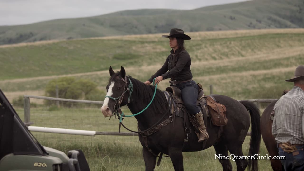 A woman dressed in cowboy attire rides a horse through a vast, open field under a clear blue sky.